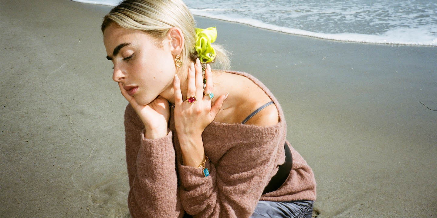 Photo of a model kneeling in the sand wearing an off-shoulder Rachel Comey sweater and wearing Mondo Mondo rings.