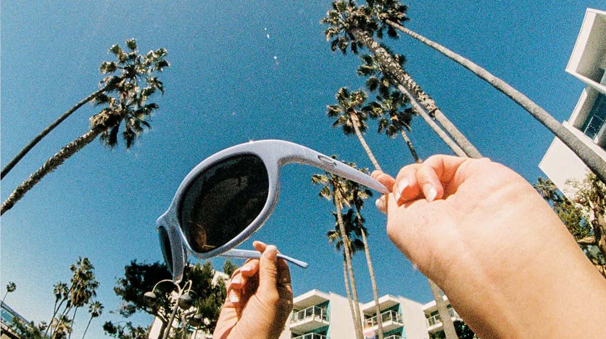 A person holding up a pair of light grey Crap sunglasses against a blue sky filled with palm trees.