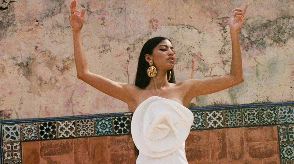 Photograph of a model wearing a sculptural white strapless dress with her arms raised in the air.