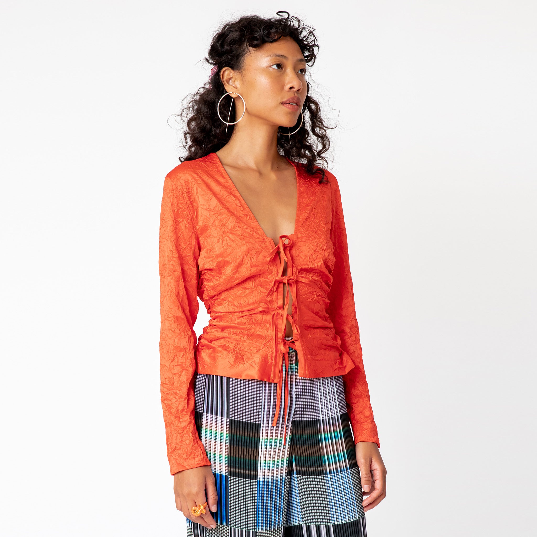 A model looks to the side while wearing a tomato red, V-necked crinkled blouse with 4 bow ties along the front.