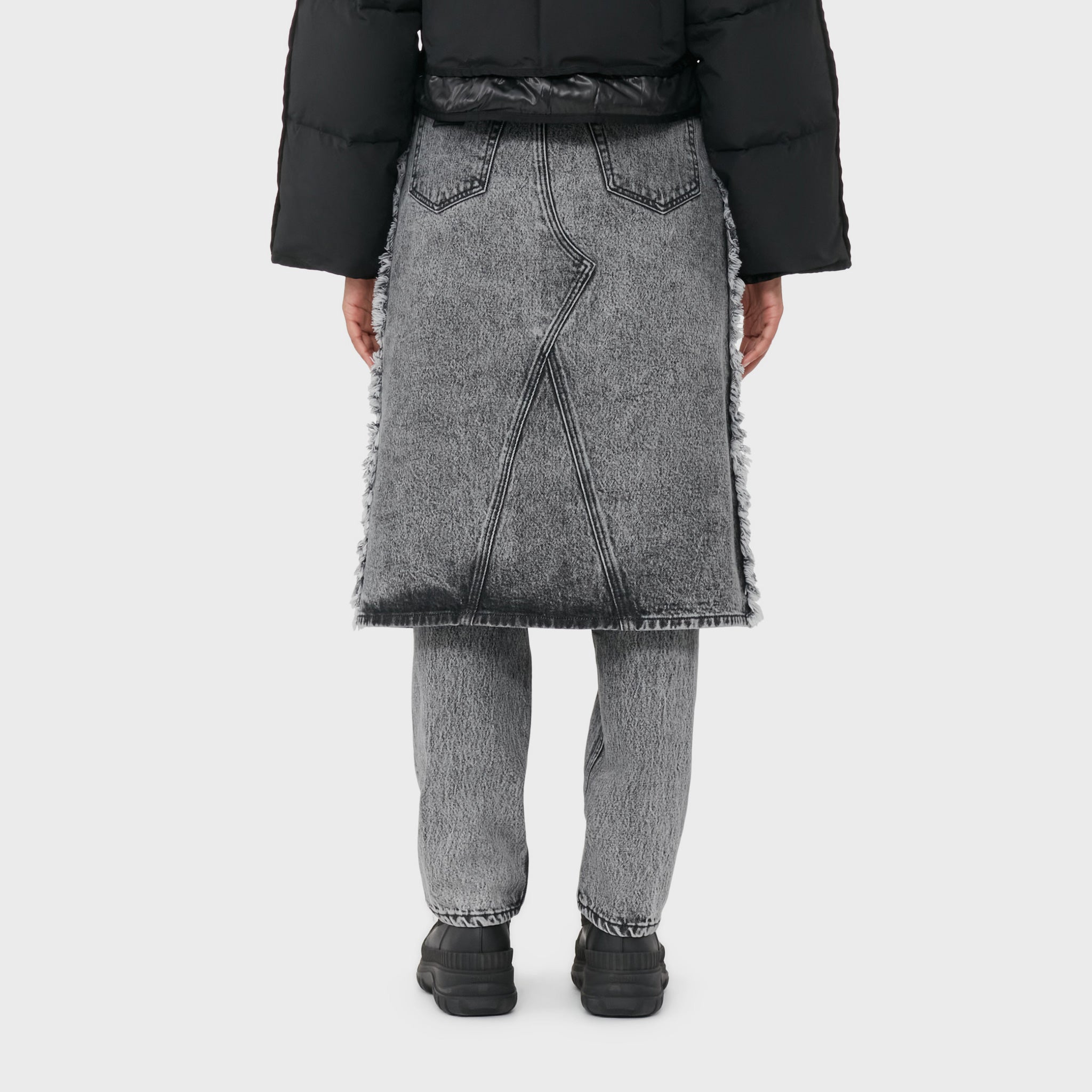 A model wears the acid washed black denim jeans with attached midi skirt with black boots - back view.