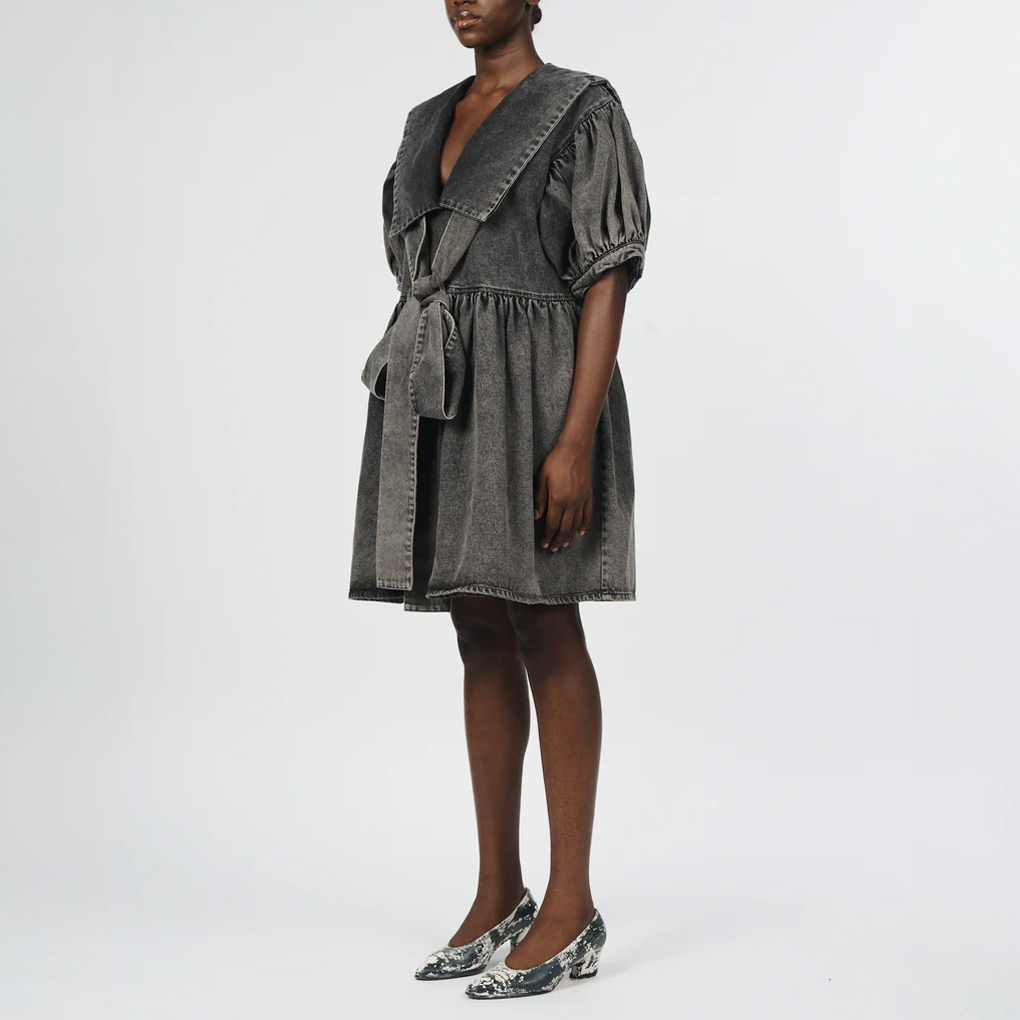 Full body photo of model wearing a denim black acid wash dress with a large collar and bow detail in the front.