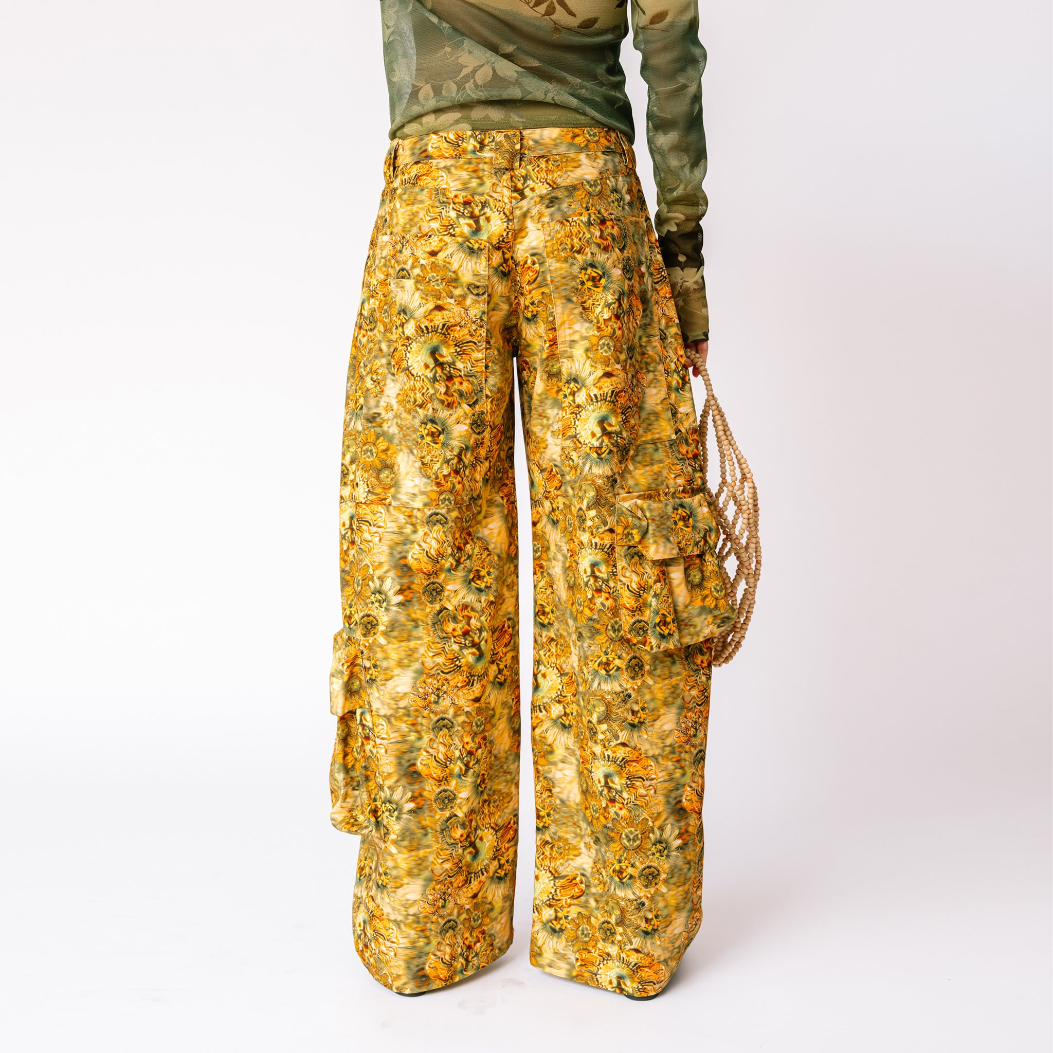 A model wears the brown and yellow floral printed Lawn Cargo Pants by Collina Strada with a mesh green long sleeved top - back view.