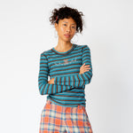 A model has her arms crossed while wearing the blue and brown striped Cardio Crew by Collina Strada featuring rhinestones on the chest in the shape of a teddy bear and swirls.