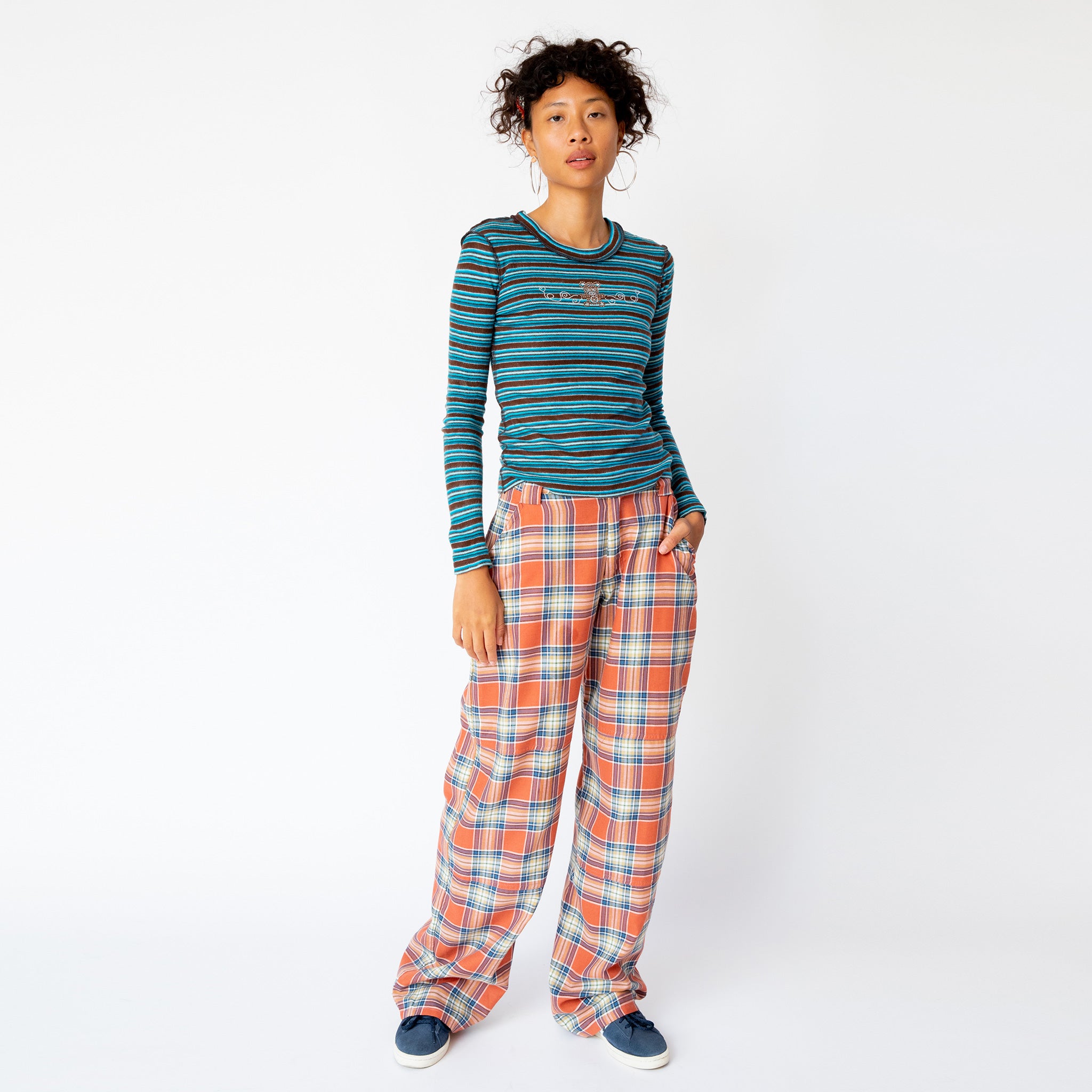 Full outfit view of the blue and brown striped Cardio Crew by Collina Strada featuring rhinestones on the chest in the shape of a teddy bear and swirls, paired with orange and white plaid wide leg pants..