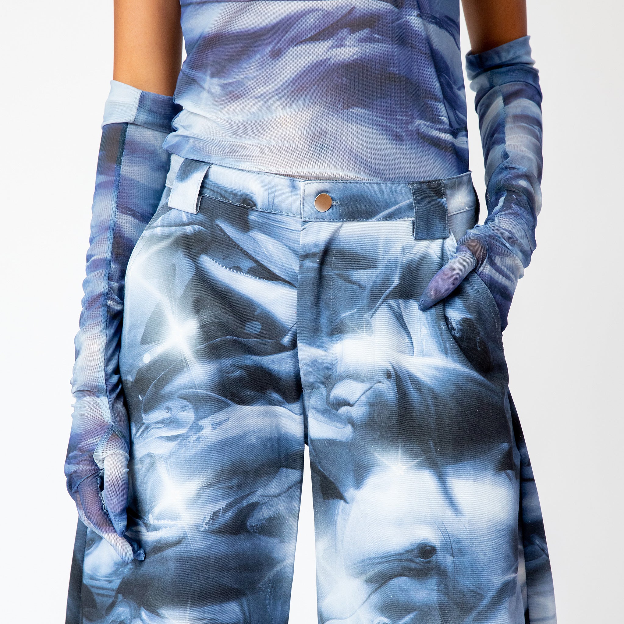 A model wears the blue and white graphic dolphin printed, wide leg Stomp Pant by Collina Strada with matching mesh top and gloves - detailed close up.