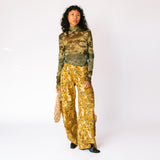 A model wears the long sleeved mesh Thumbtastic Top in an ombre green print with floral and leaves silhouettes - full outfit view paired with yellow and brown floral cargo pants.