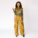 A model wears the long sleeved mesh Thumbtastic Top in an ombre green print with floral and leaves silhouettes - full outfit view paired with yellow and brown floral cargo pants.