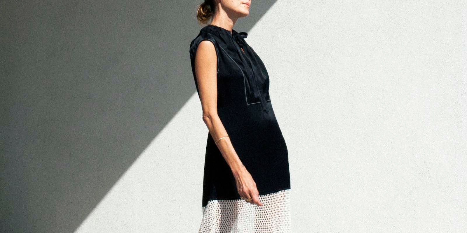 Model stands in the sun against white wall wearing a black sleeveless dress with white crochet bottom. 