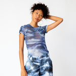 A model holds one hand up to her head while wearing the blue and white printed mesh Arc Top by Collina Strada with matching cargo pants.