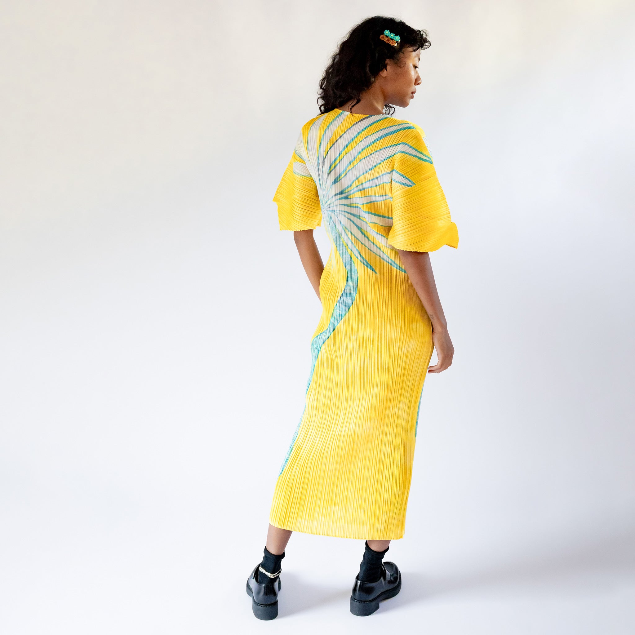 A model wears the full length yellow pleated Puff Sleeve Dress with a large graphic print of a desert plant, paired with black socks and loafers, back view.