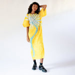 A model holds one hand to her head as she wears the full length yellow pleated Puff Sleeve Dress with a large graphic print of a desert plant, paired with black socks and loafers.