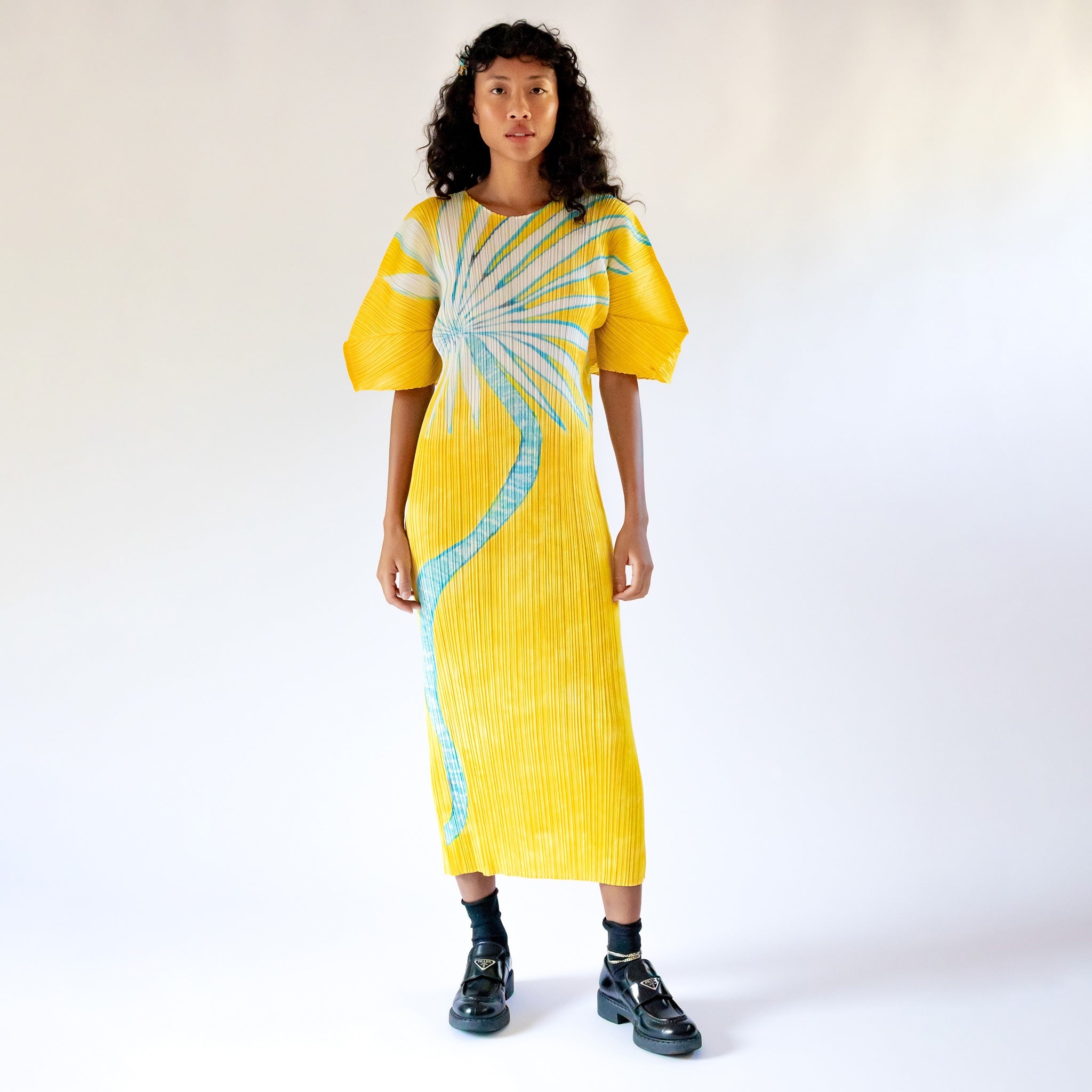 A model wears the full length yellow pleated Puff Sleeve Dress with a large graphic print of a desert plant, paired with black socks and loafers.