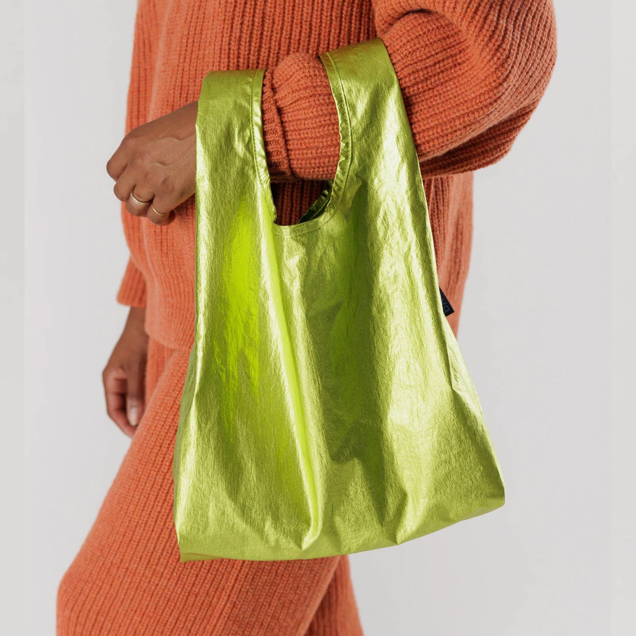 Smaller version of BAGGU's classic reusable nylon tote, in a metallic green color, shown on the arm of a woman wearing orange knits.