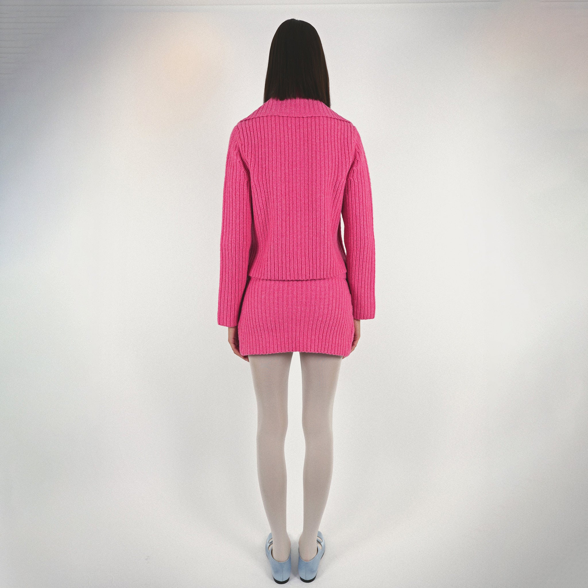 Back full body photo of model wearing a pink knit set featuring a button up sweater and a mini skirt.