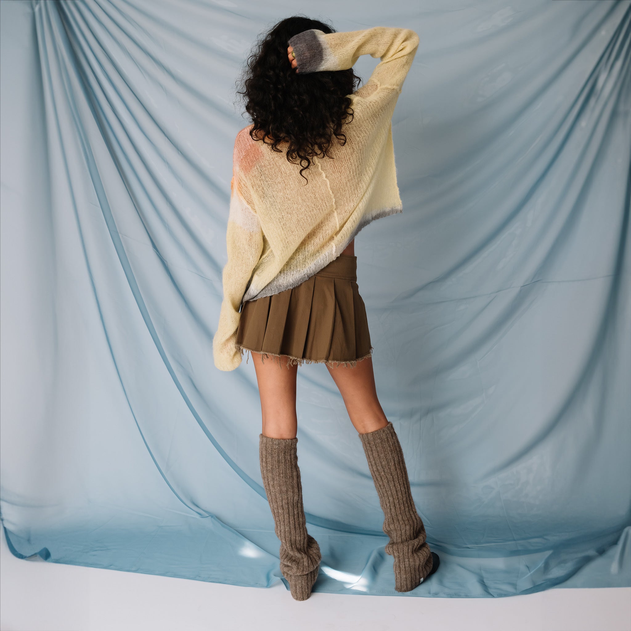 A model wears the Misc Etc belted pleated mini skirt in khaki, paired with a pale yellow sweater, leg warmers and black loafers, backview.