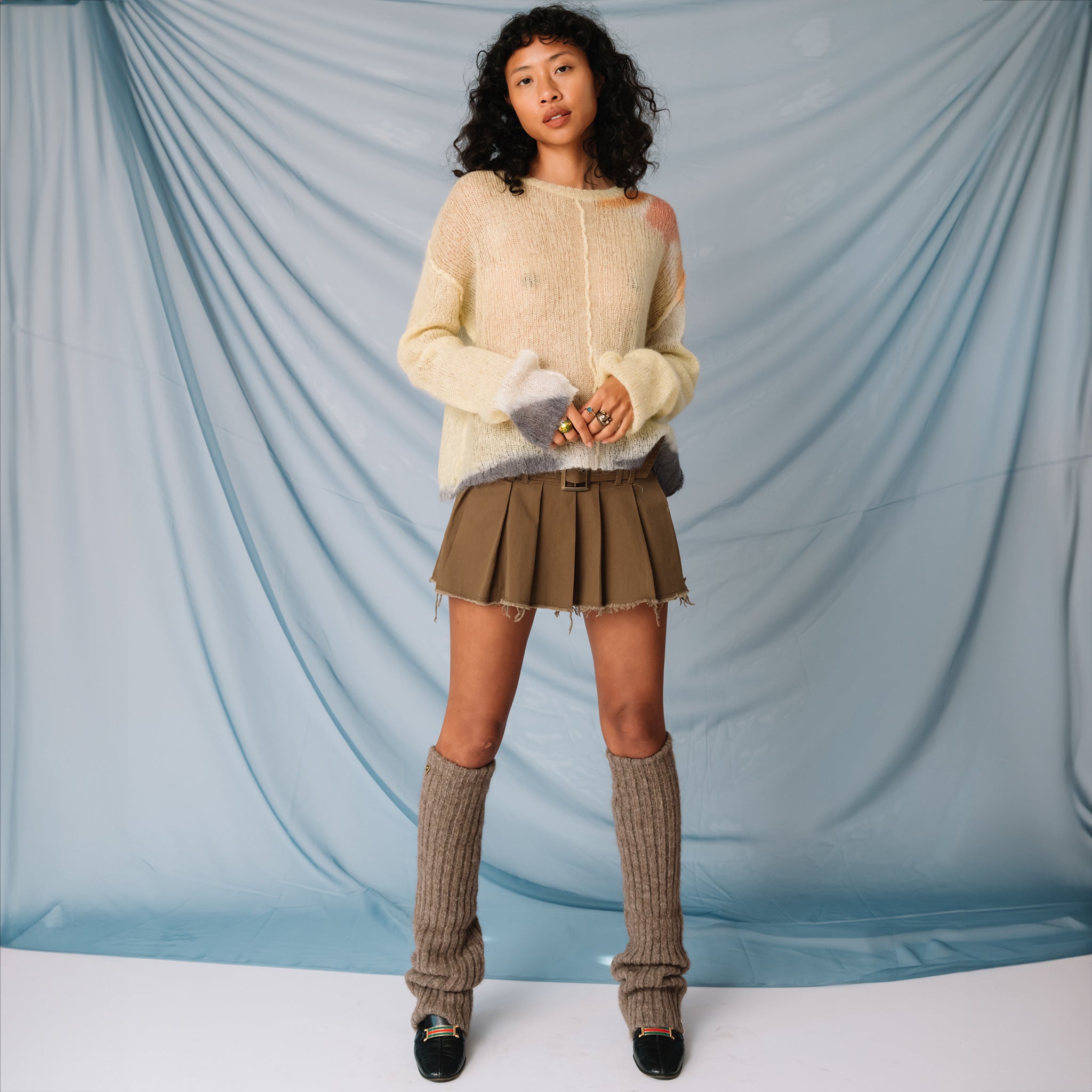 A model wears the Misc Etc belted pleated mini skirt in khaki, paired with a pale yellow sweater, leg warmers and black loafers, full outfit view.