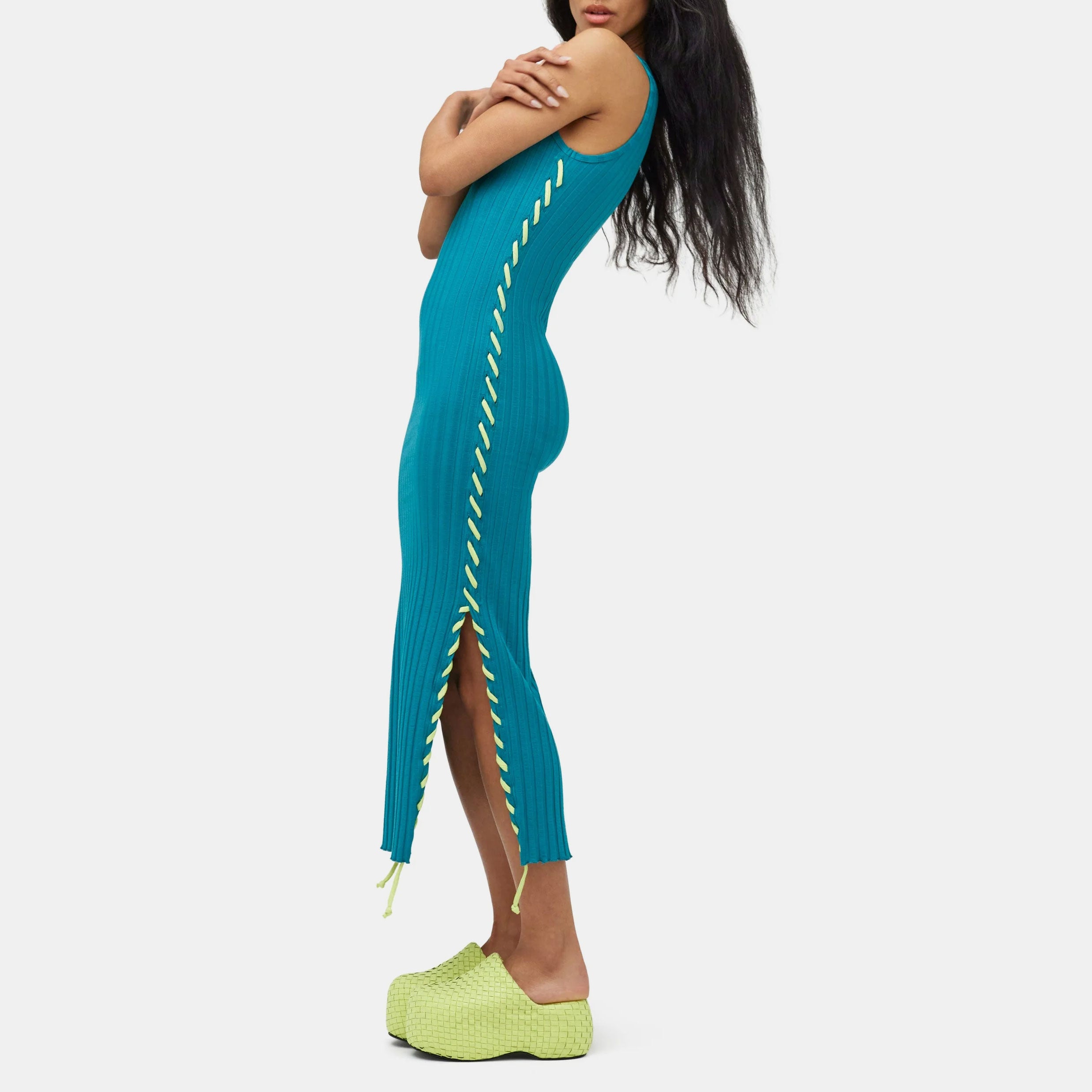 Full body photo of model wearing the teal green ribbed Blix Dress, a sleeveless ribbed dress with fluorescent green oversized stitching detail along the sides - left side view.