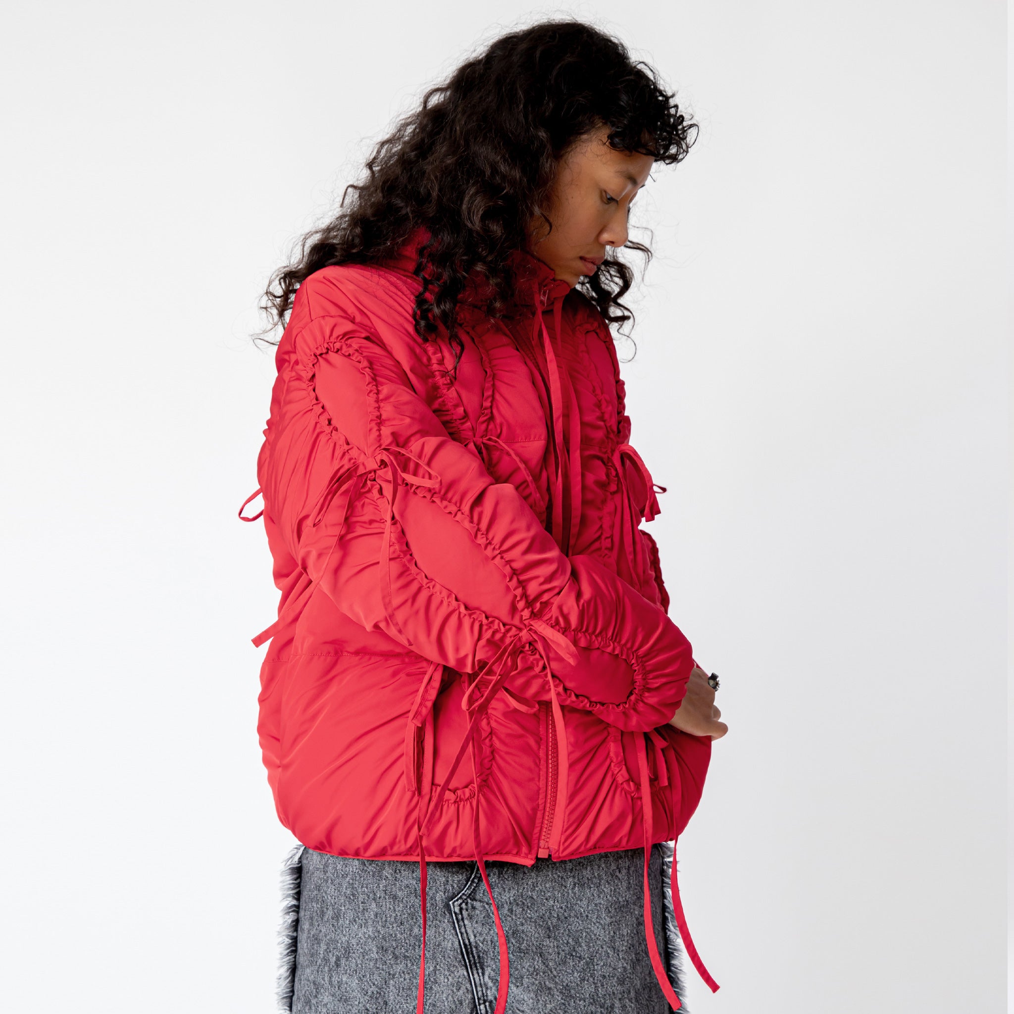 Side half body detail photo of model wearing a red puffer jacket with ruching bow details. 