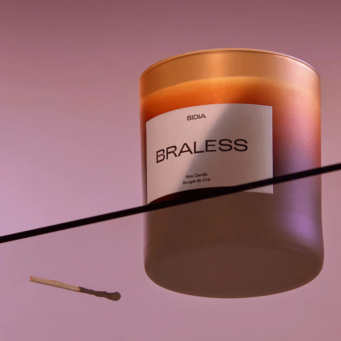 Photo of the Candle - Braless sitting on top of a clear tabletop with a small match next to the product. 