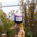 A Nirvana candle with frosted purple glass is sat upon a gnarled tree root with wildflowers and a lake in the background.