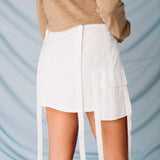 Back view of the white cargo mini skirt with dangling white straps, paired with a light brown cardigan.