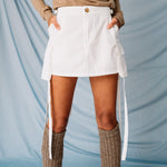 Detailed front photo of a model wearing the white cargo mini skirt with dangling white straps, paired with a light brown cardigan and leg warmers.