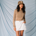 A model looks to the side while wearing the white cargo mini skirt with dangling white straps, paired with a light brown cardigan.