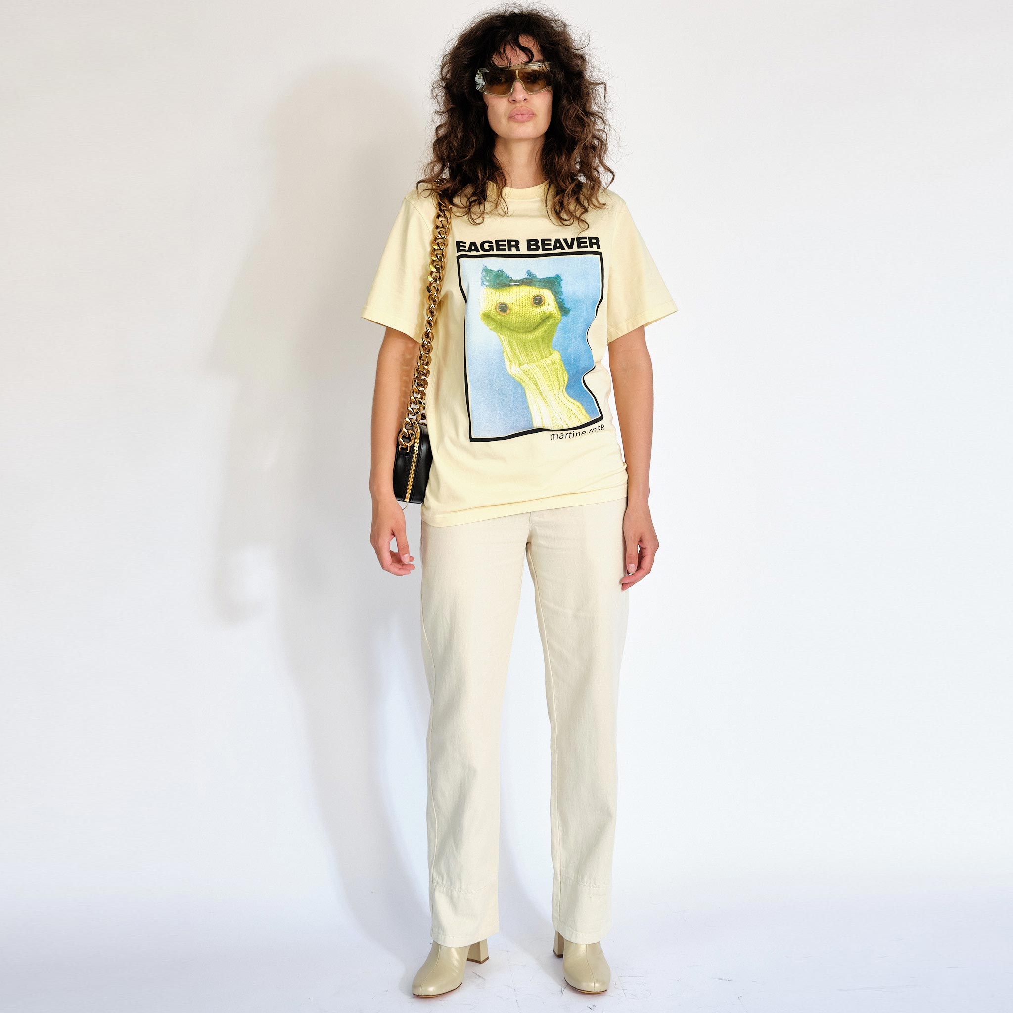 A model wears the light yellow Martine Rose t-shirt featuring a sock puppet graphic and the words Eager Beaver on the front chest - full outfit view.
