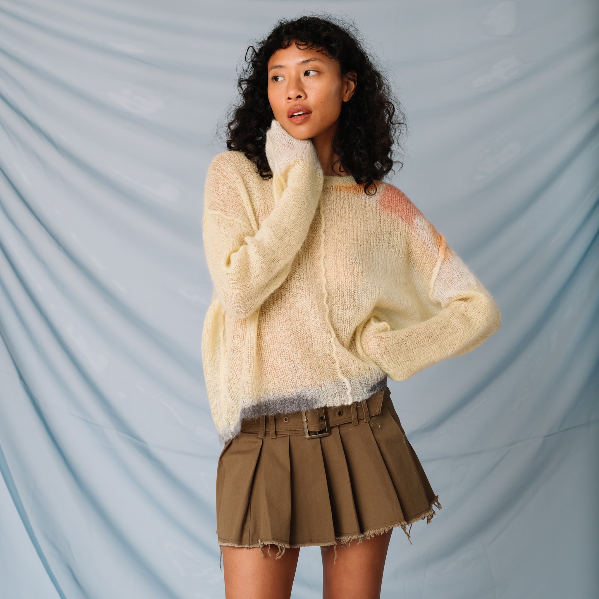 A model looks to the side while wearing the pale yellow Composition sweater by Eckhaus Latta, paired with a pleated mini skirt, front view.