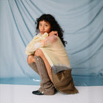 A model crouches down while wearing the pale yellow Composition sweater by Eckhaus Latta paired with a pleated mini skirt, leg warmers and black loafers.