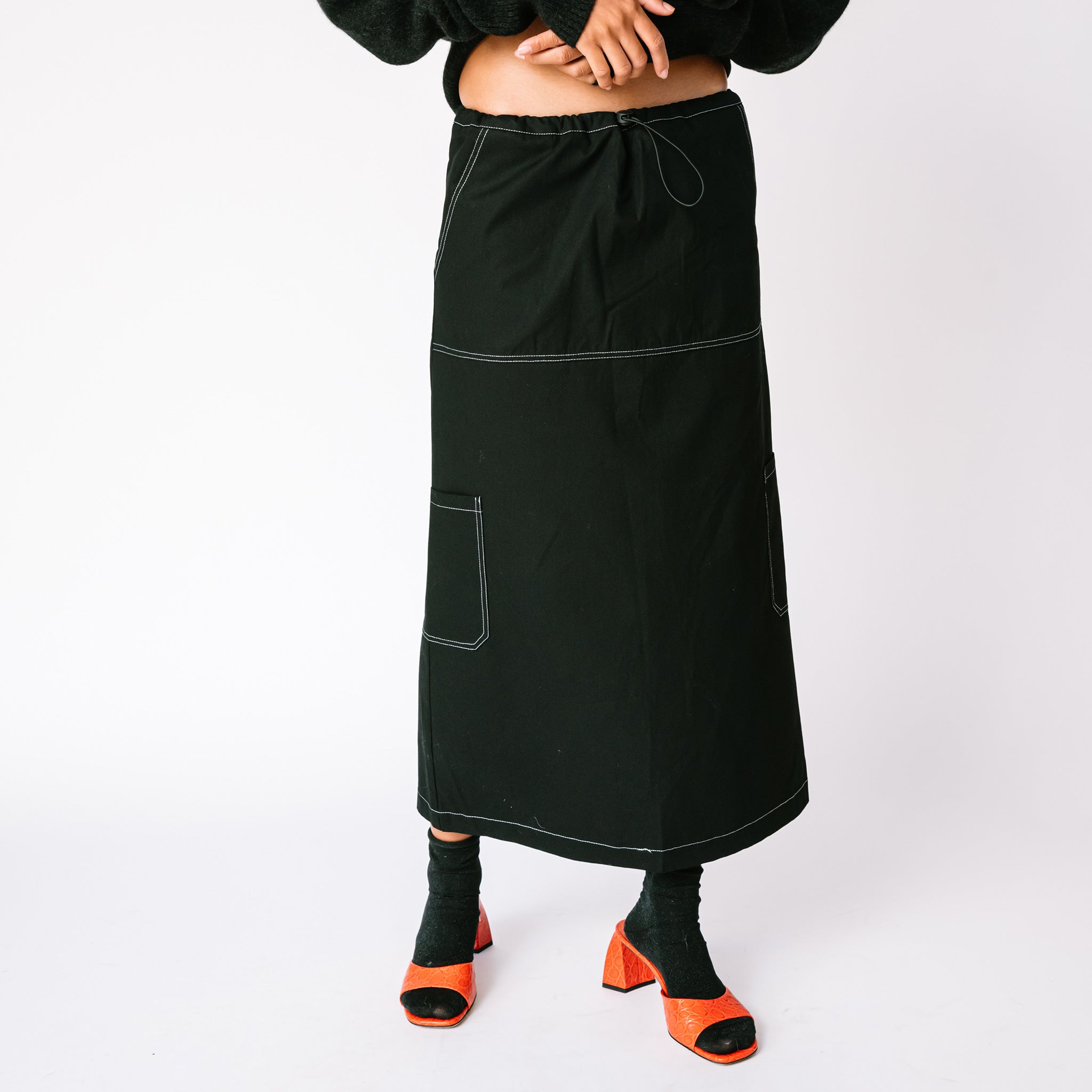 A model wears the Misc Etc long Contrast Stitch Cargo Skirt in black - paired with black socks and red heels.