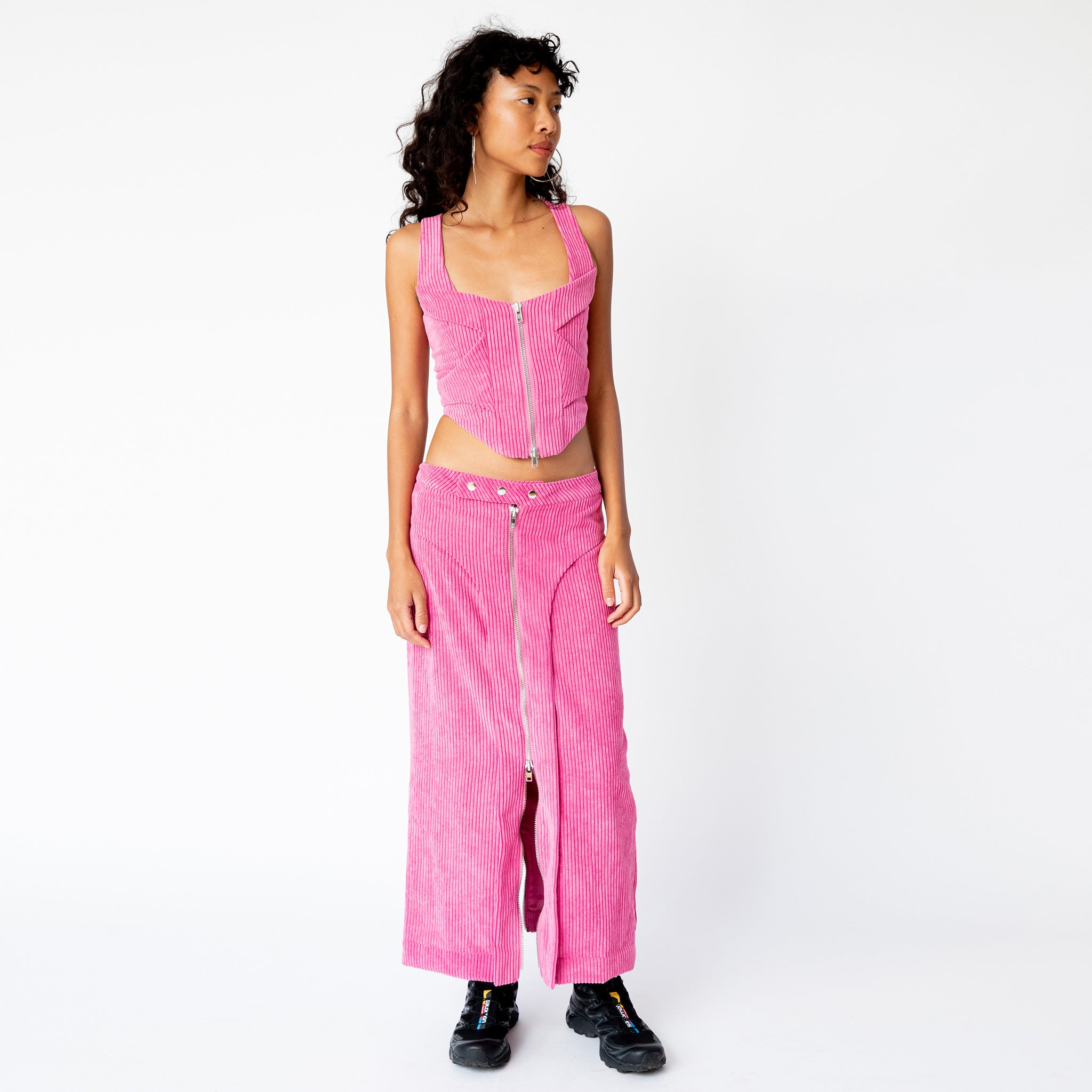 A model looks to the side while wearing the pink corduroy Cord Bustier by Eckhaus Latta, with silver front zipper and wide shoulder straps, paired here with the matching full length corduroy zip skirt.