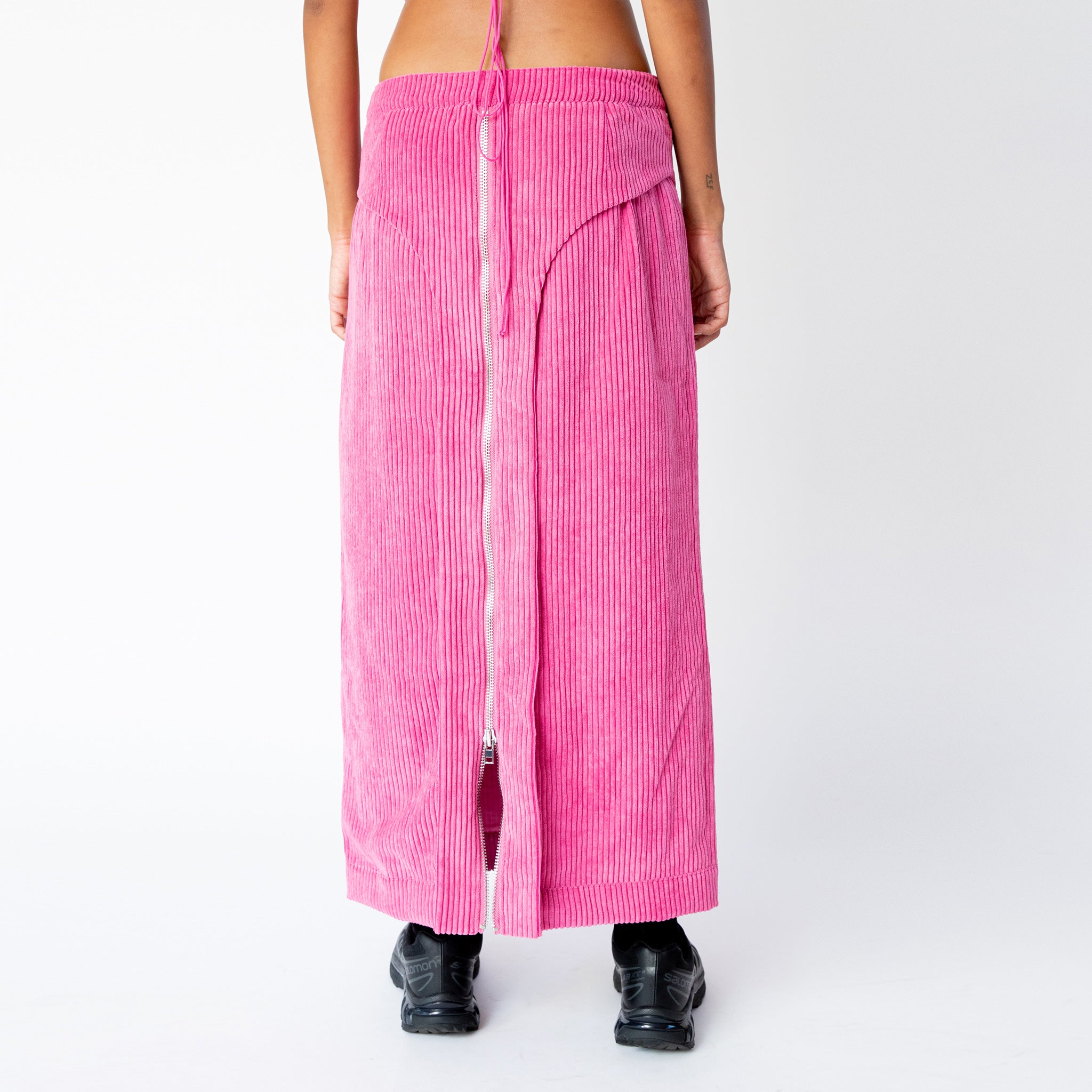 A model wears the full length pink corduroy Cord Zip Skirt by Eckhaus Latta, with full length silver front zipper and 3 front waistband button snaps, paired here with black sneakers - back view.