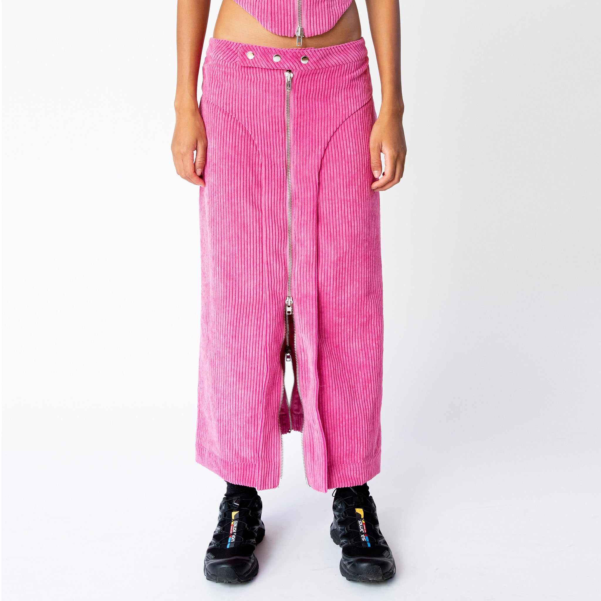 A model wears the full length pink corduroy Cord Zip Skirt by Eckhaus Latta, with full length silver front zipper and 3 front waistband button snaps, paired here with black sneakers.