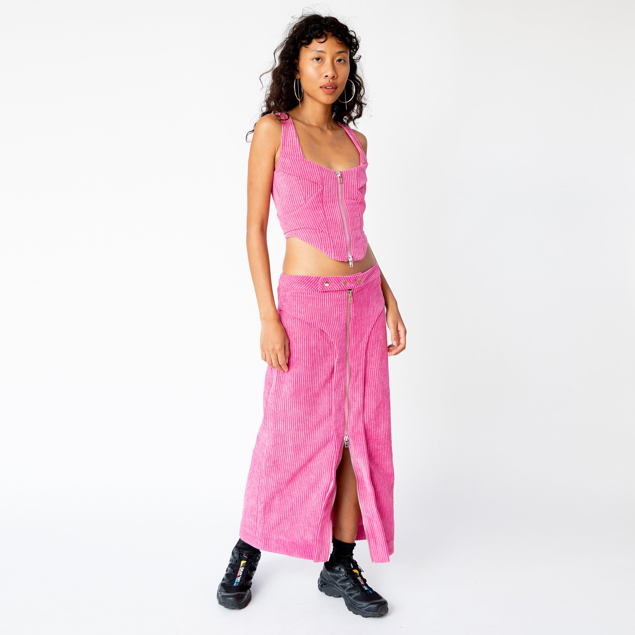 A model wears the full length pink corduroy Cord Zip Skirt by Eckhaus Latta, with full length silver front zipper and 3 front waistband button snaps, paired here with the matching full length corduroy zip up bustier and black sneakers.