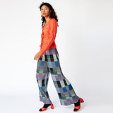 A model walks to the side while wearing a tomato red, V-necked crinkled blouse with 4 bow ties along the front paired with graphic pleated pants.