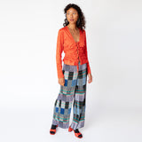 A model walks to the side while wearing a tomato red, V-necked crinkled blouse with 4 bow ties along the front paired with graphic pleated pants.