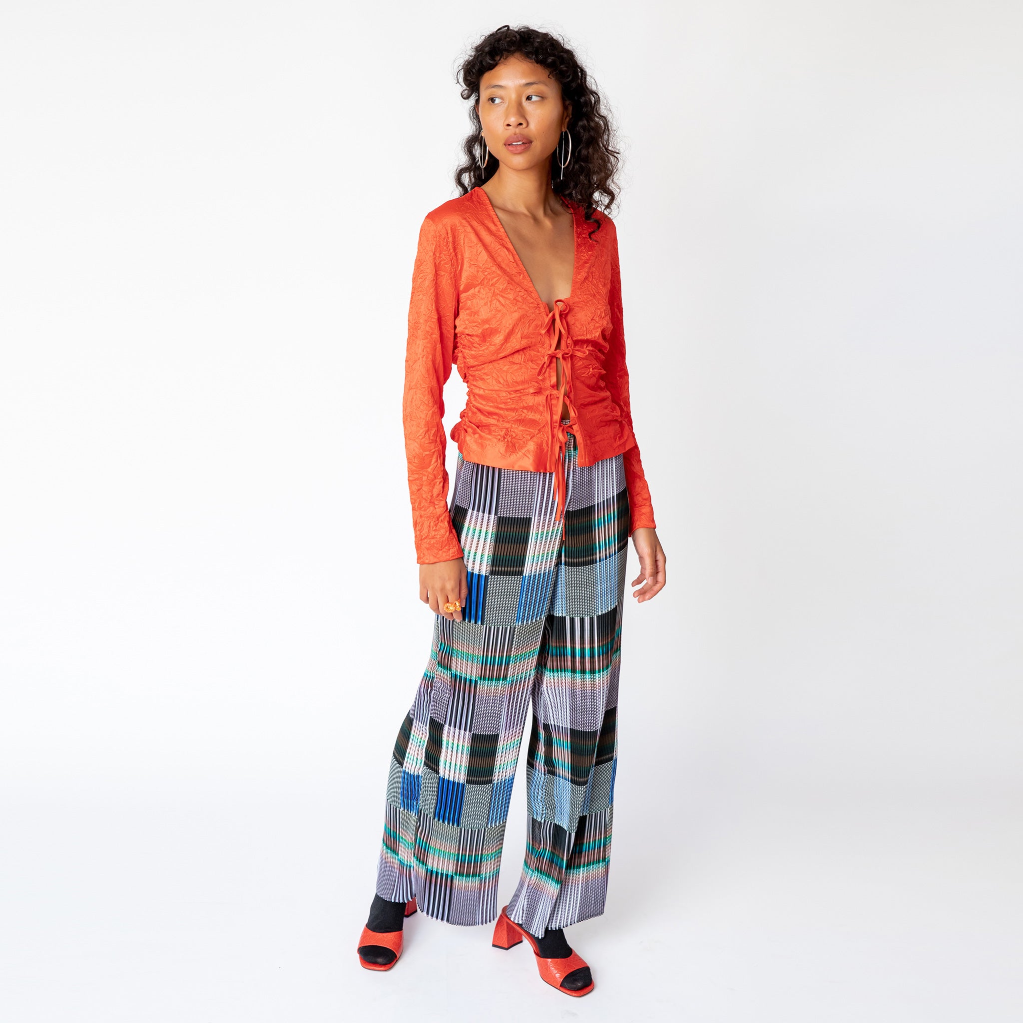 A model walks to the side while wearing a tomato red, V-necked crinkled blouse with 4 bow ties along the front paired with graphic pleated pants.