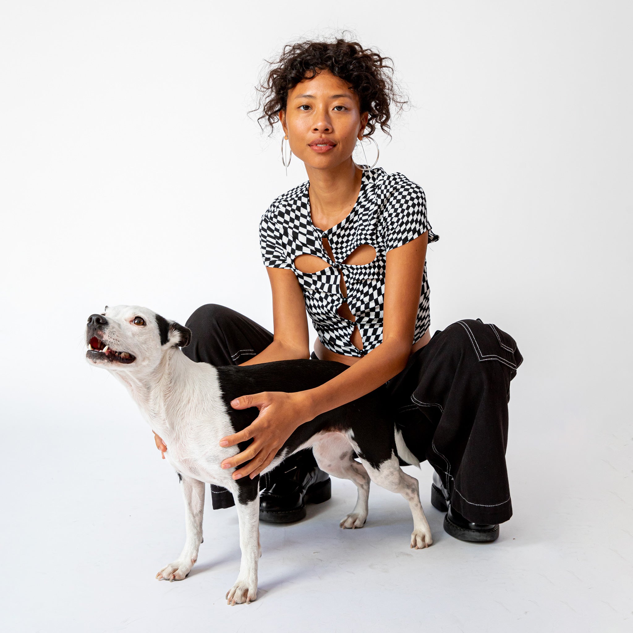 A model crouches down to pet a small white dog while wearing the black and white checkered cut out tee by Misc Etc, paired with black cargo pants.