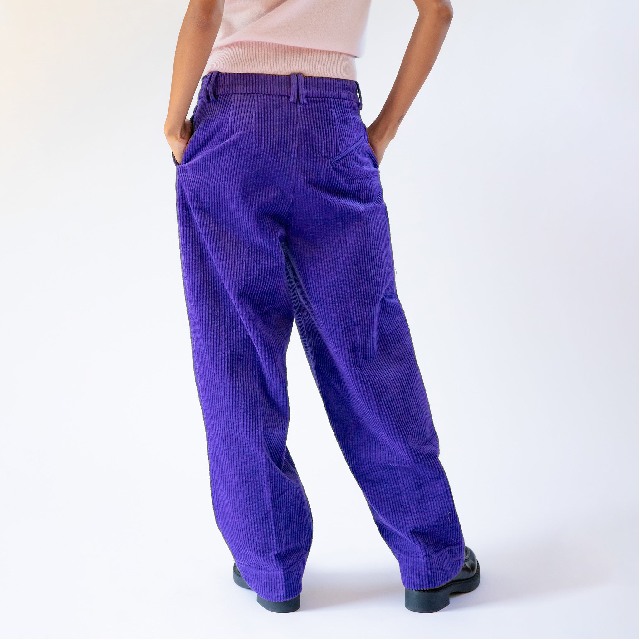 Back photo of a model wearing the GANNI relaxed corduroy pants in purple, paired with a simple grey vest and black loafers.