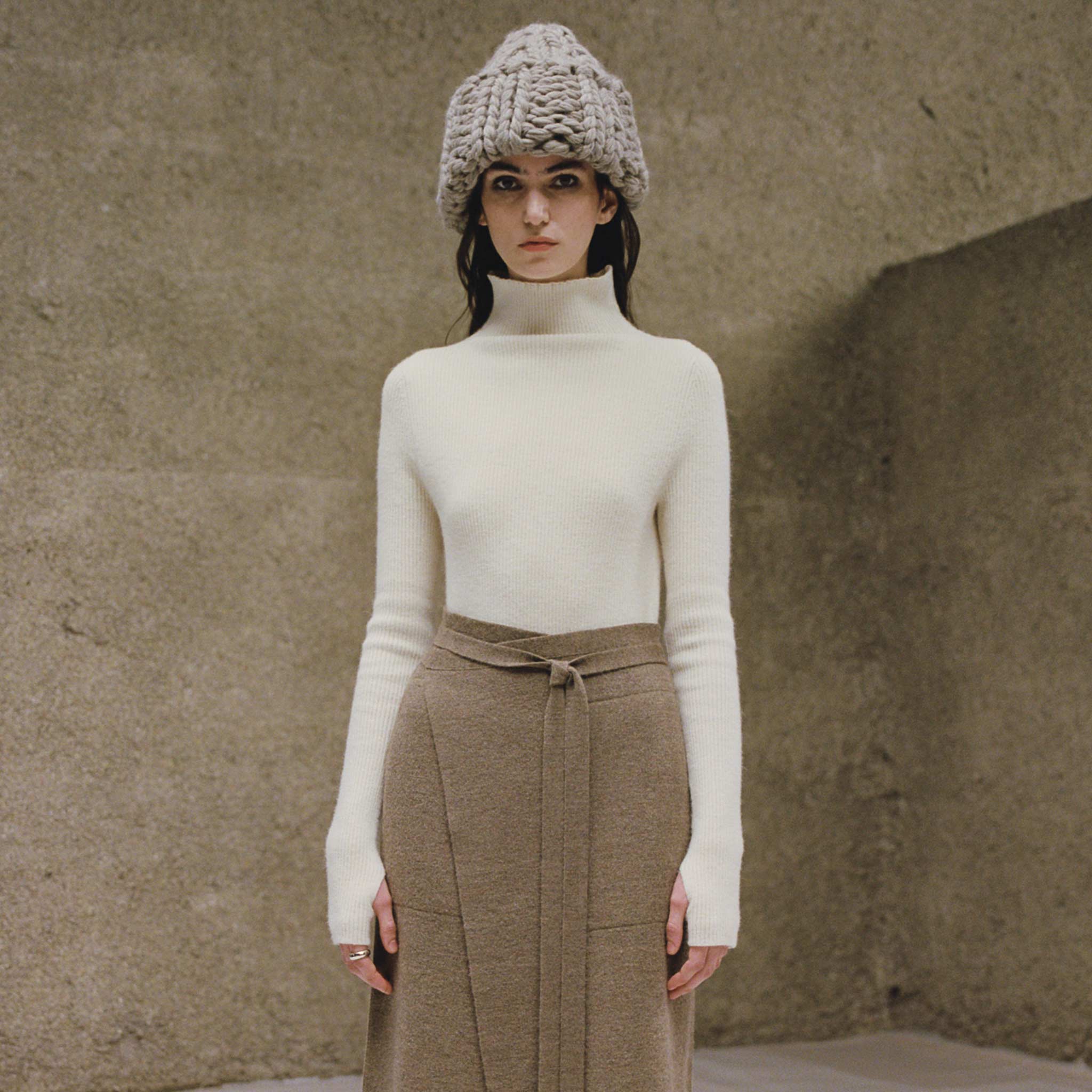 A lookbook image of a model wearing the ribbed white mockneck knit top with a wide high collar mockneck and thumb hole details at the sleeves, front view.
