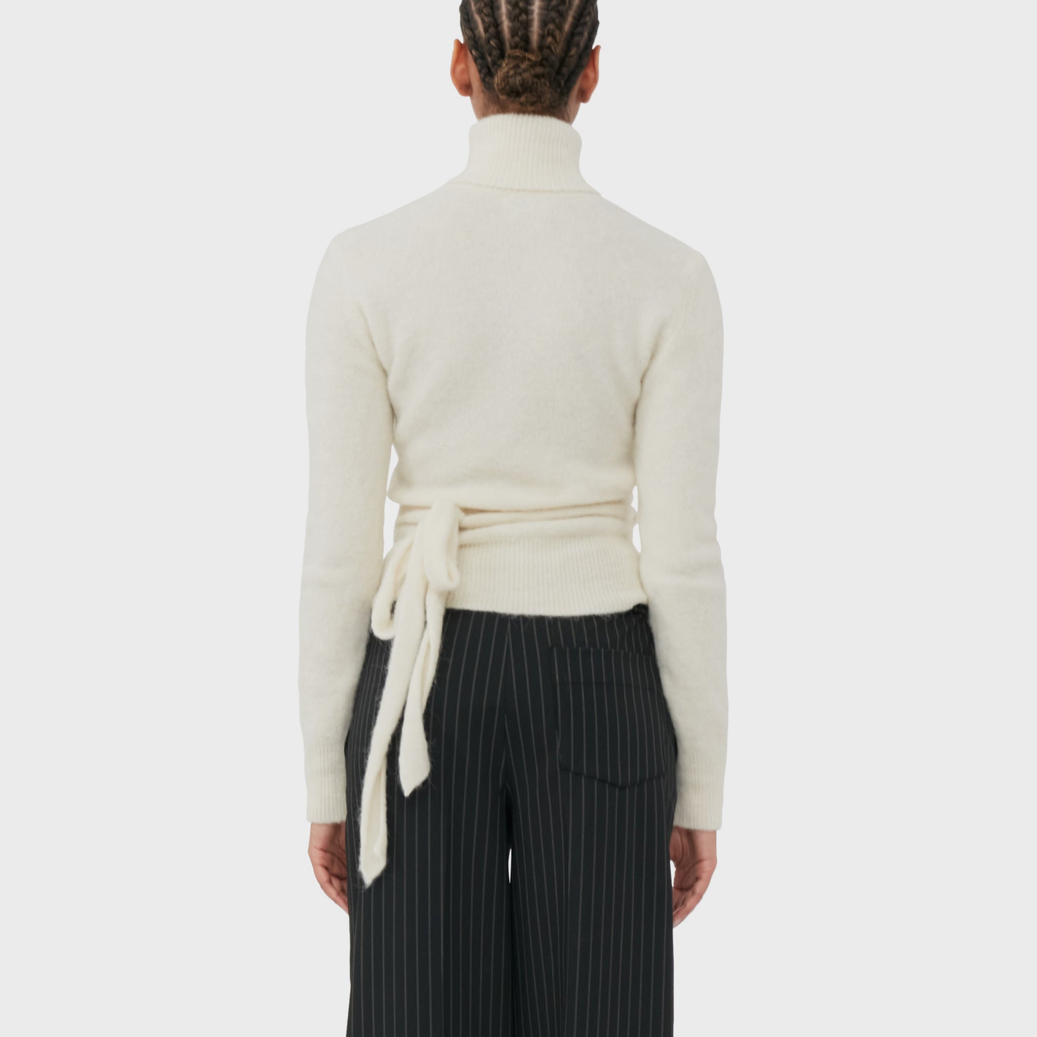 A model wears the soft wrapped white turtleneck sweater with cutout details from GANNI - back view showing waist tie.