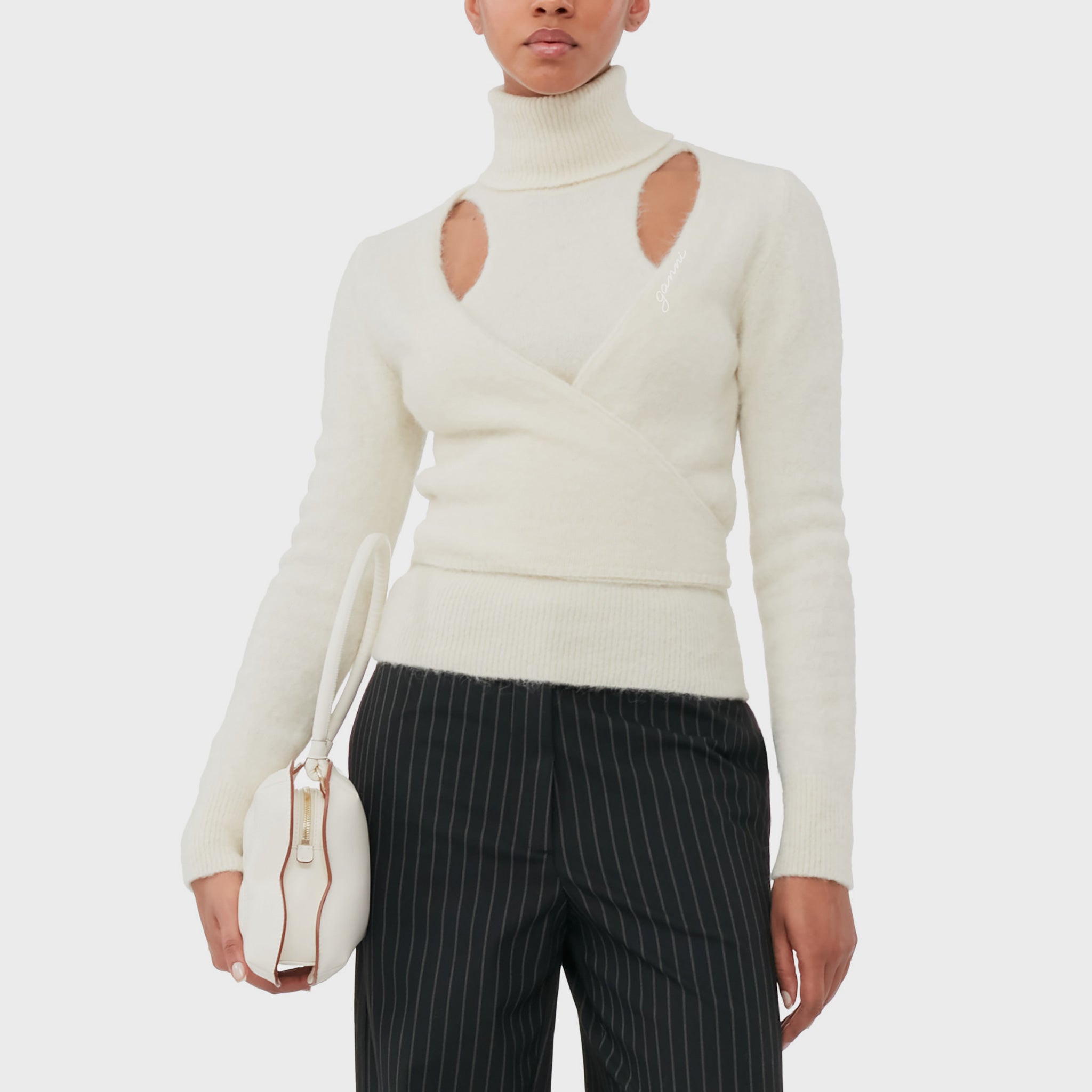 A model wears the soft wrapped white turtleneck sweater with cutout details from GANNI.