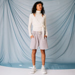 A model looks to the side while wearing a soft-looking white wrap sweater by GANNI with clavicle cut outs, paired with pale lavender sweat shorts.