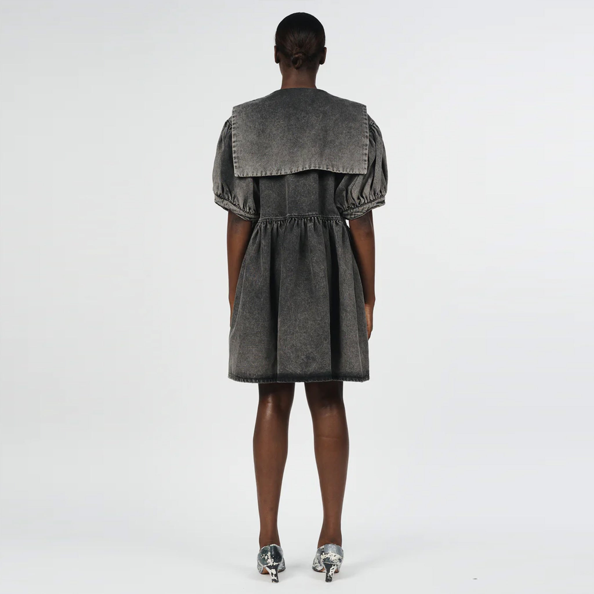 Back full body photo of model wearing a mini denim acid wash dress with a large collar and bow detail. 