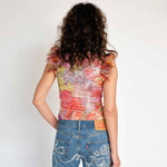 A model wears the Dachi top - a multicolored stretch lace tank top with pink feathers around the arm holes - back view.