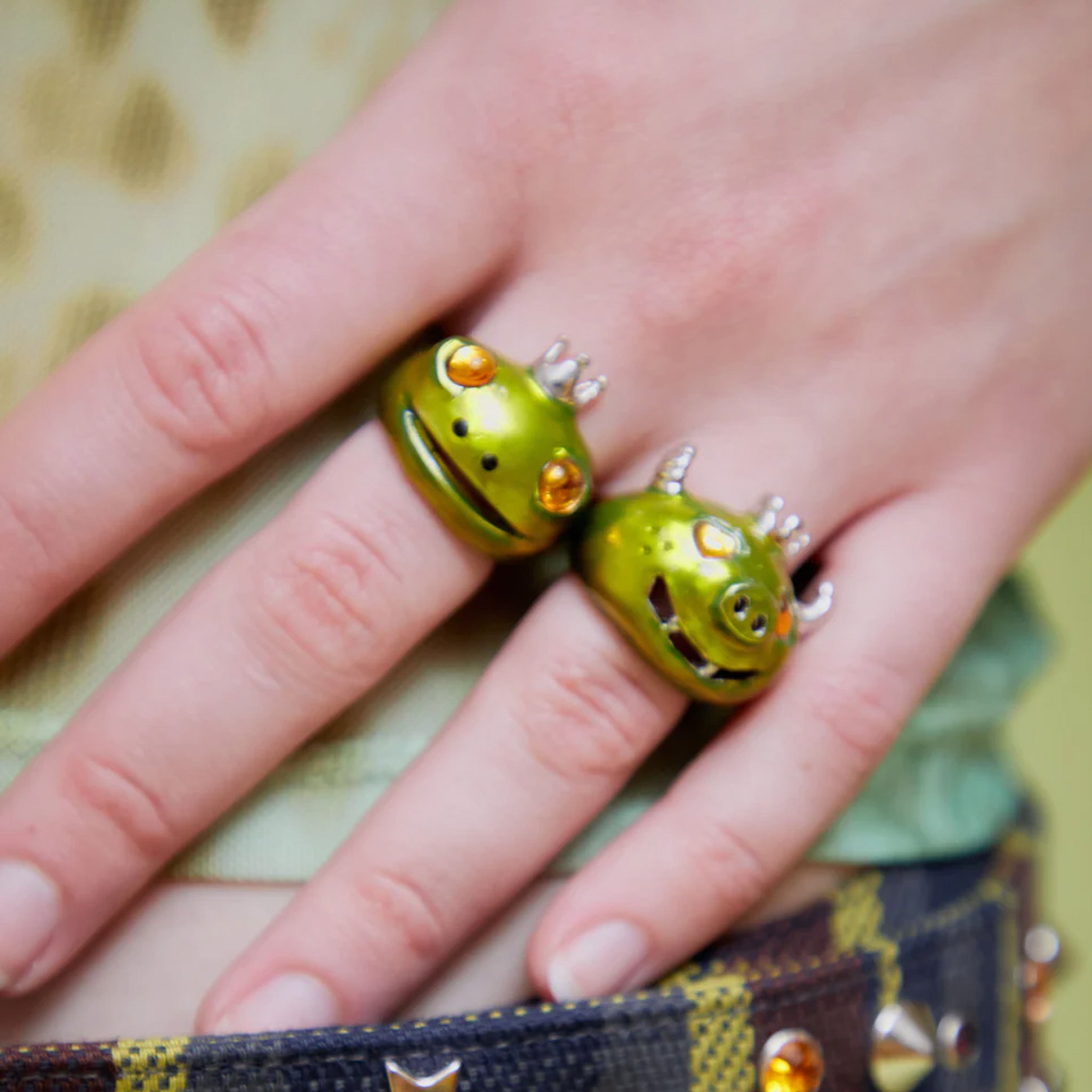 A model wears The Frog Prince Ring, a recycled pewter ring shaped like a frog's head wearing a tiny silver crown, hand painted in lime metallic green with golden glass eyes.