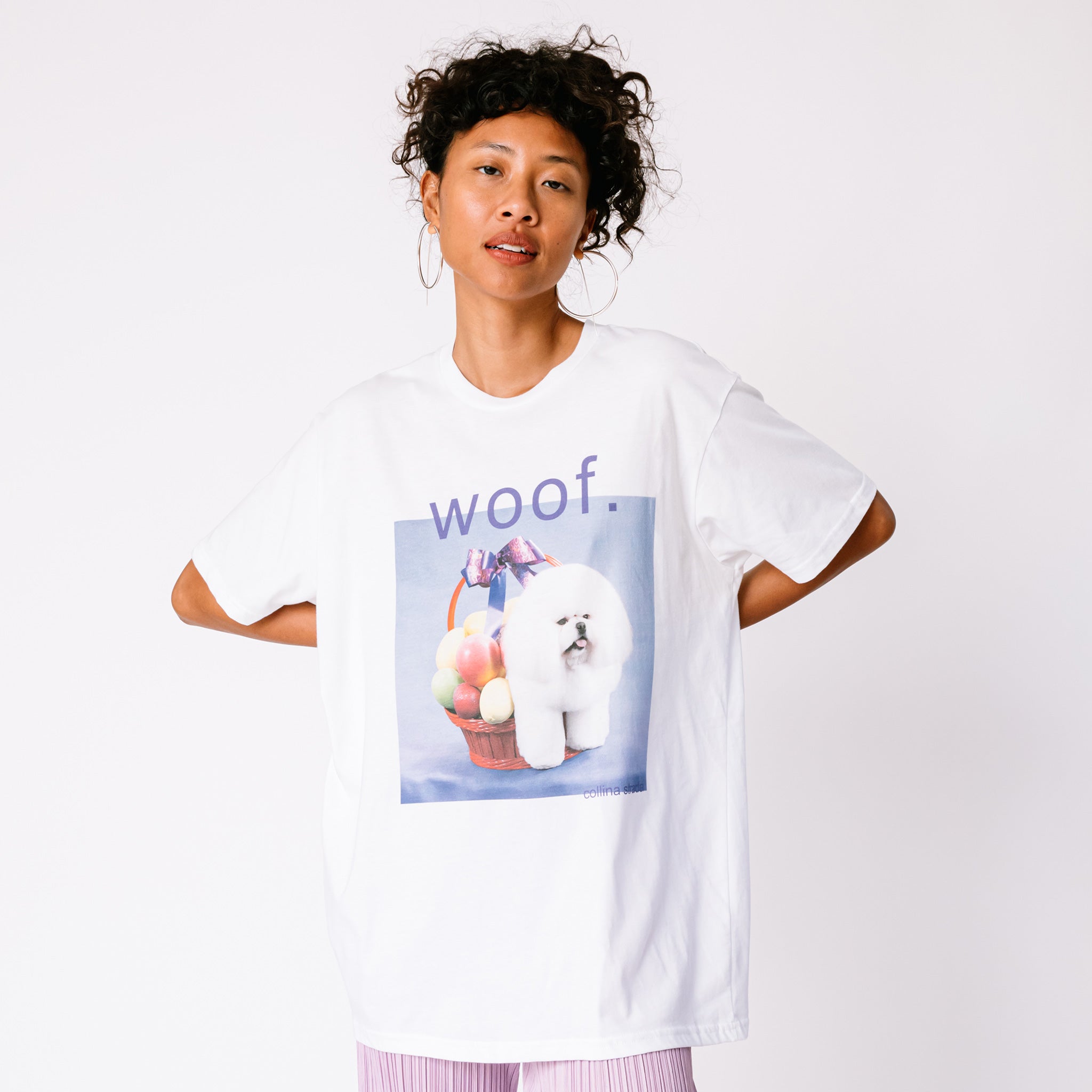 A model wears the white graphic tee by Collina Strada featuring a photo of a white fluffy dog with its rear legs hidden in a fruit basket and the word woof printed askew above.