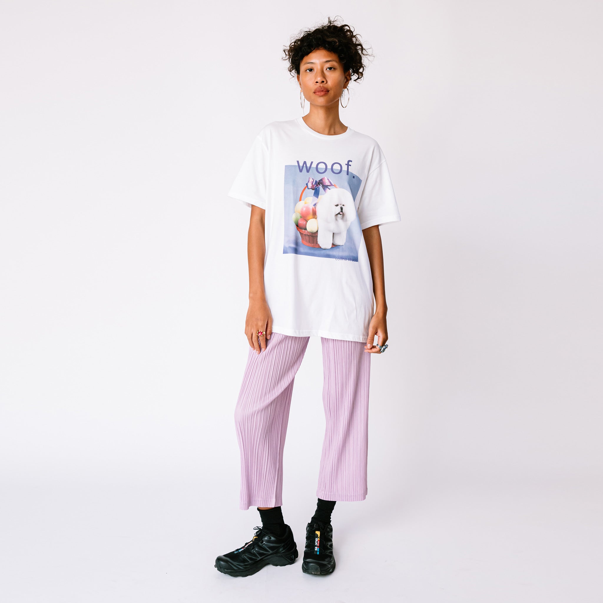 A model wears the white graphic tee by Collina Strada featuring a photo of a white fluffy dog with its rear legs hidden in a fruit basket and the word woof printed askew above, paired with pleated pants.